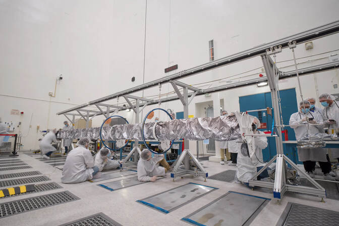 Workers crouch and crawl underneath the boom, which resembles a TV cable tower wrapped in aluminum foil. The boom is hanging from a track raised off the ground by 6 tripod-like stands.