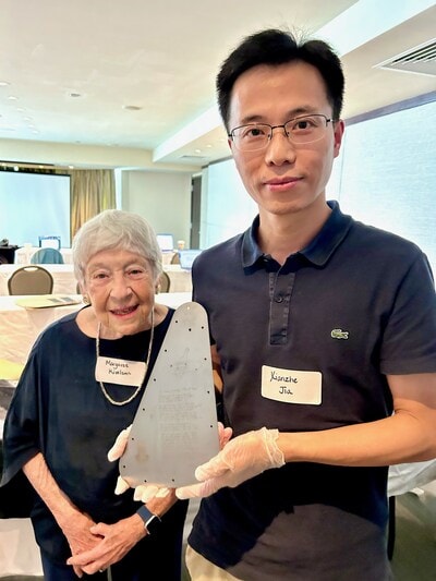 A man and a woman stand in a conference room. The man is holding a triangular metal plate with gloved hands.