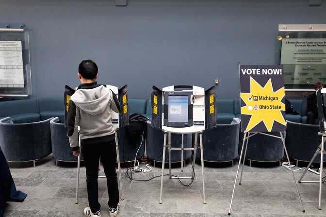 A sign next to two voting machines reads "Vote now!" with a checked box for Michigan and an empty box for Ohio State. The screen of one machine is visible but unreadable, while a voter blocks the view of the other.