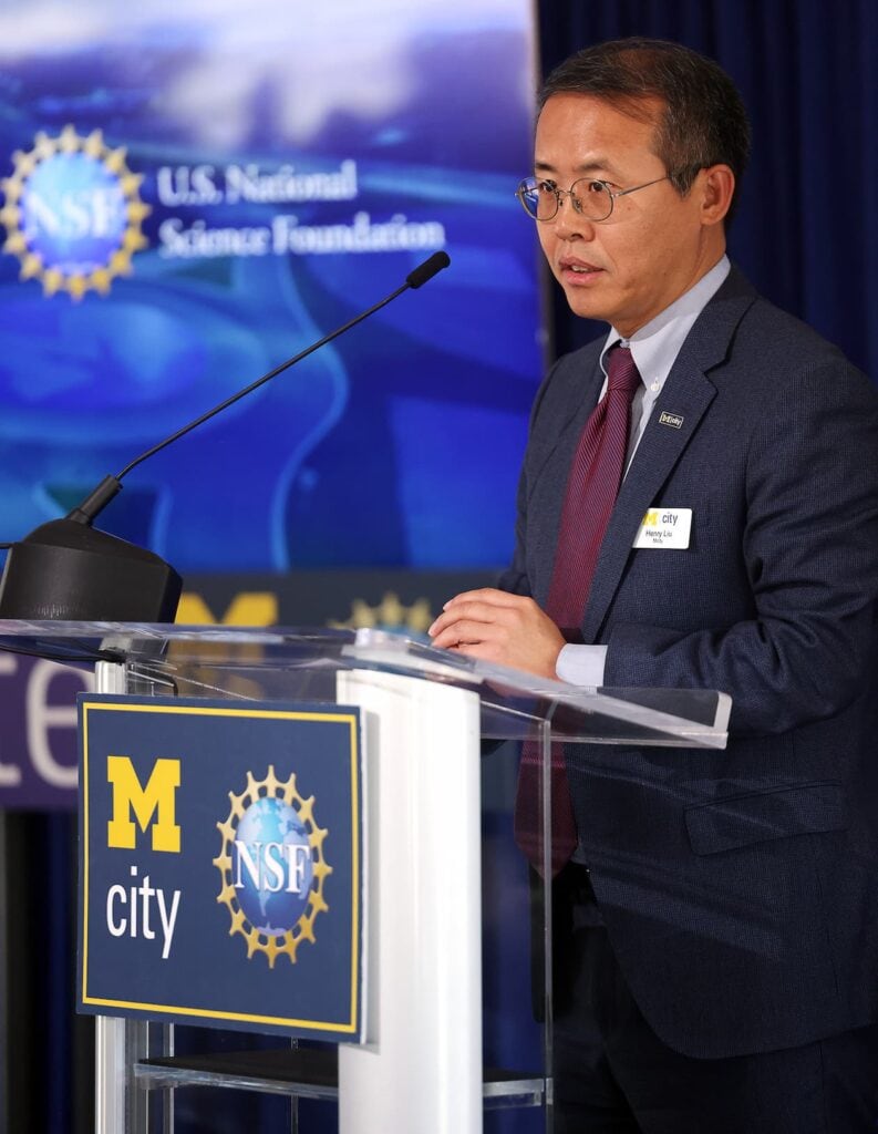 A man, wearing a blue suit and maroon tie, speaks at a podium.