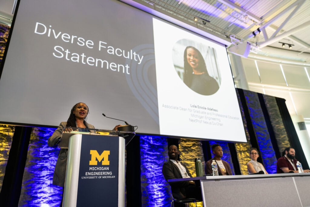 Lola stands at a podium. Behind her on the screen reads "Diverse faculty statement"