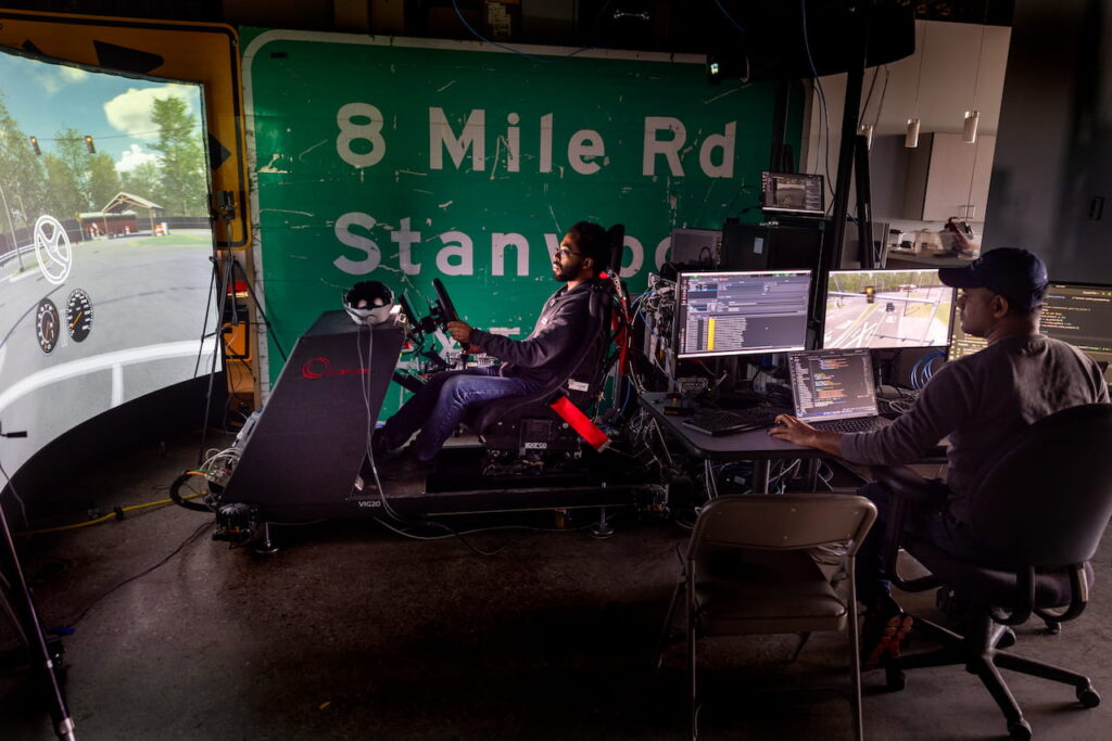 One man sits, reclined as a driver would, with a steering wheel and dashboard set up immediately in front of him, along with a large screen further away that gives a view from the driver's perspective on a remote controlled car. To the right, sits another man at a desk behind a bank of computer screens.