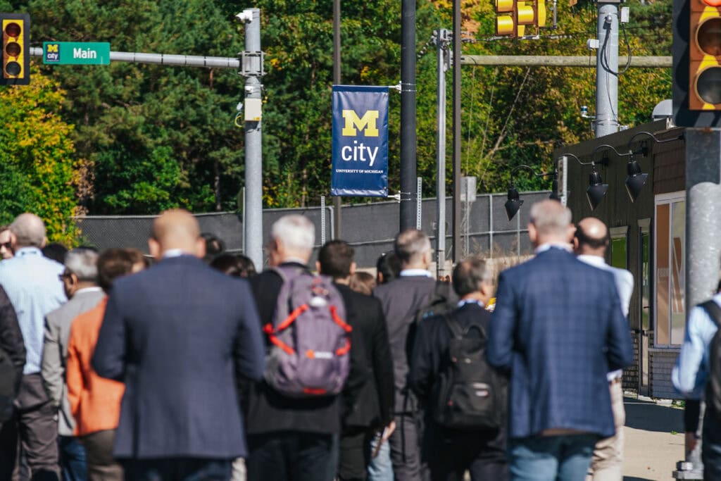 A group of a dozen people are photographed from the back as they walk toward Mcity Test Facility. Above them on a street banner there is a Mcity logo.