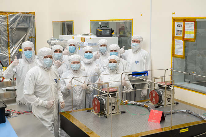 Ten engineers wearing white clean suits and blue medical masks standar around an instrument that resembles two metal boxes. The boxes rest on a waist-height table and a red drum-like structure protrudes from a face on each box.