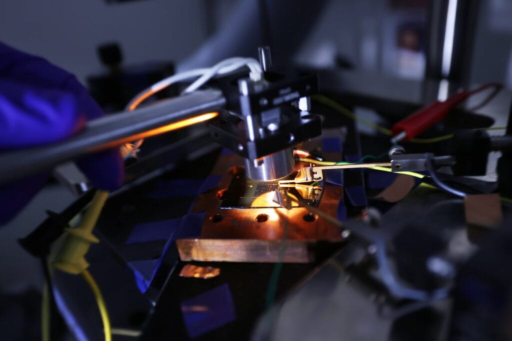 A steel arm holds a black box over a thin piece of gold, which is secured to a lab bench and connected to several wires. A metal cylinder protrudes from the black box and points toward a gray square on the gold sheet, which is the photovoltaic cell's semiconductor material. Orange light glows from the cylinder and reflects off the gold sheet.