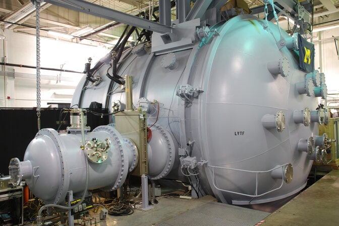 The vacuum chamber is a pale gray cylinder with rounded ends and various protrusions, about 3 times as tall as a person and 5 times as long. A block M flag hangs high on the visible end.