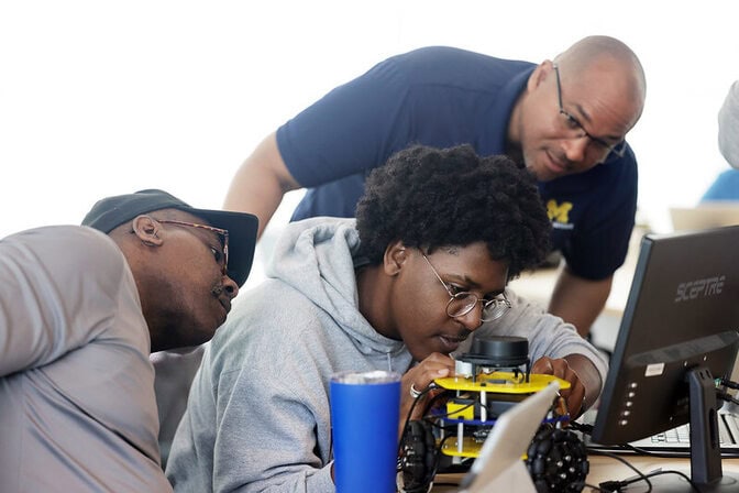 Three people engaged with a small robotic vehicle on a desk table.