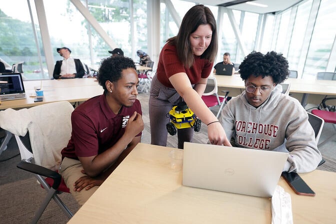 Three people gathered around a laptop, two seated and one standing, in a bright room with others sitting at tables behind them. The person in the middle is pointing at the laptop screen while holding a robotic device, while the other two individuals look at the laptop screen.