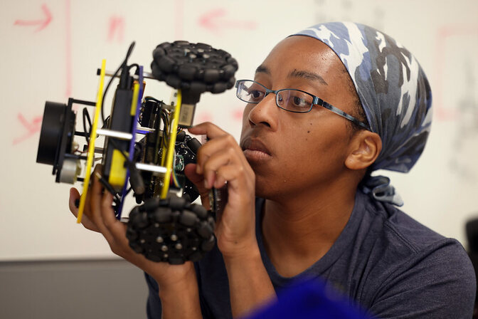 Close-up of a person examining the bottom of an MBot, a small, yellow robotic device with larger, rigid, black wheels. 