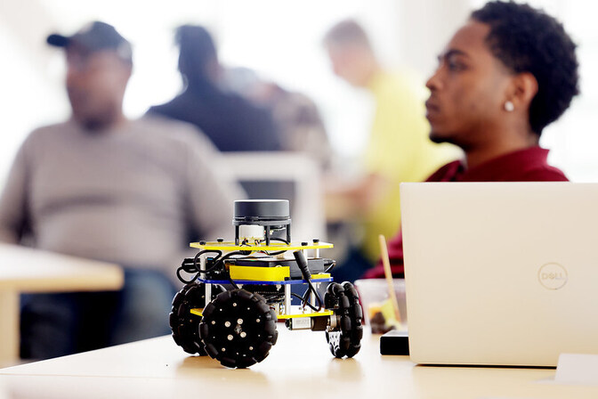 A small, yellow robotic device with larger, rigid, black wheels is placed on top of a desk next to a laptop. 