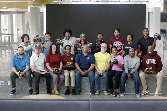 Group of thirteen people arranged in two rows sitting indoors the Ford Moto Company Robotics building, some holding robotic devices.