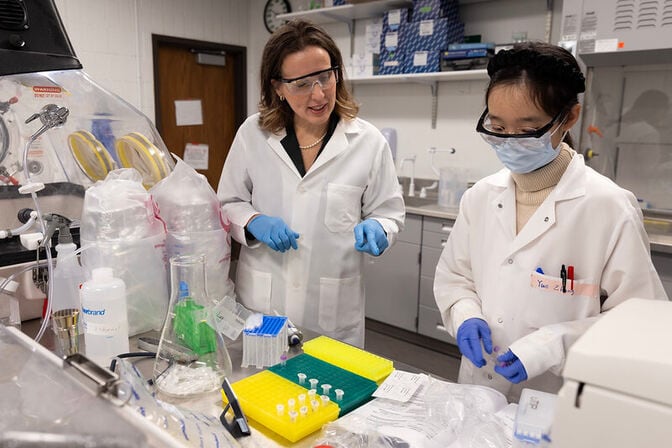 Two people in lab coats, protective eyewear and gloves look at small test tubes in a lab, with larger beakers and other lab equipment in the background.
