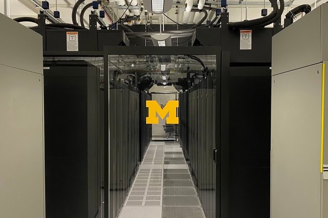 A glass door emblazoned with the U-M block M leads to a server room with black computer towers arranged in neat rows.