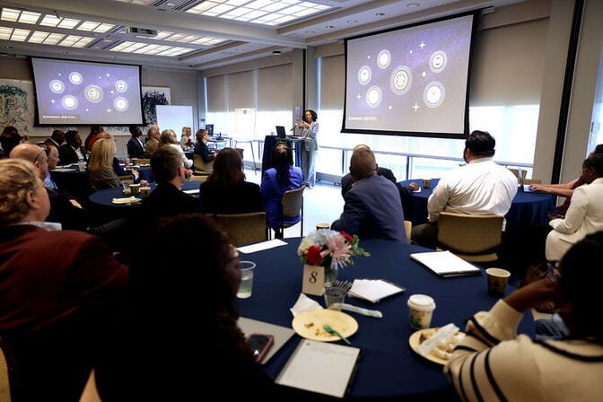   A conference room with attendees seated at tables facing a presenter and two screens displaying graphics.