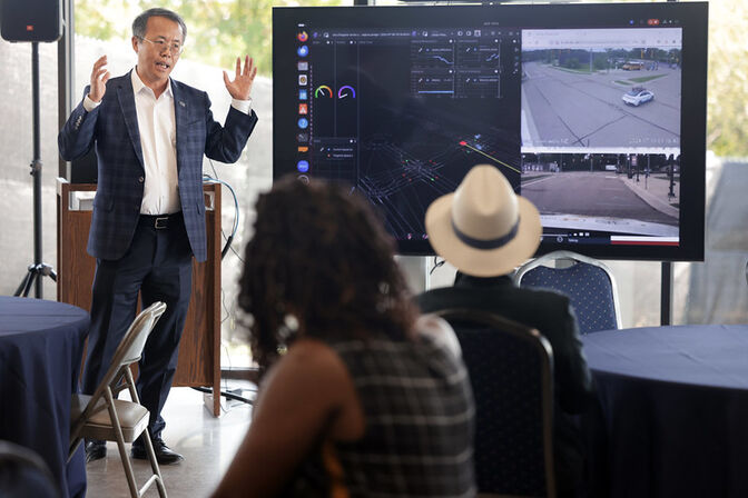 Henry Liu gestures while presenting in front of a screen displaying digital graphics and street map images, with audience members seated in the foreground.