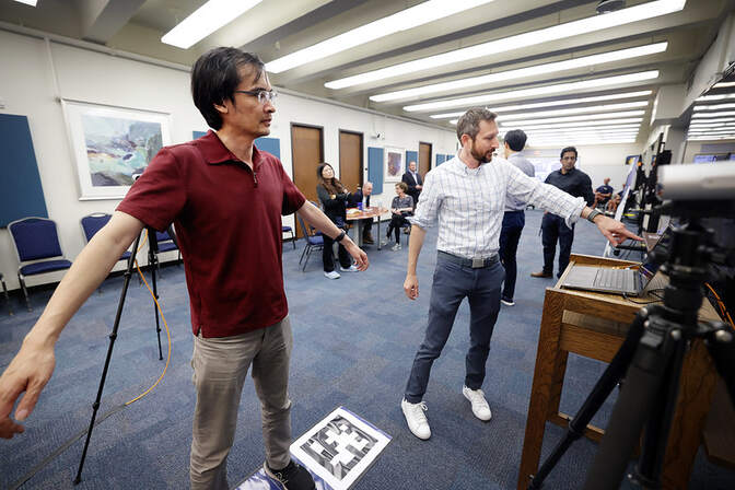 Two men are engaged in discussion over a laptop on a wooden table. One man, in a maroon polo shirt and khaki pants, stands with his arms slightly outstretched, while the other, in a white checkered shirt and jeans, points towards the screen. A black-and-white patterned square lies on the floor near them. In the background, several people sit at a long table, working on laptops or watching the interaction.