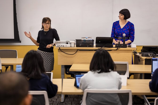 A view of a college classroom from the second row to the front podium. Two professors stand at the front of the room, one leaning and lecturing while the other looks on. Students sit at desks facing the lecturers with laptops open in front of them.