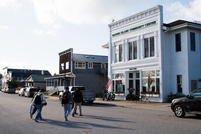 Four people cross the street toward a white, square building with two floors. Above the second-story windows, "Beaver Island Community Center" is spelled out in green letters.