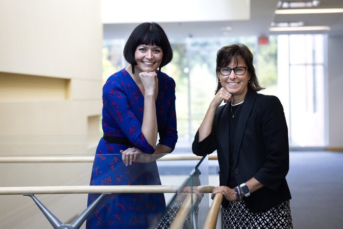 Two women pose next to each other, smiling with their face propped up on their first, in a sunny hallway of an academic building.