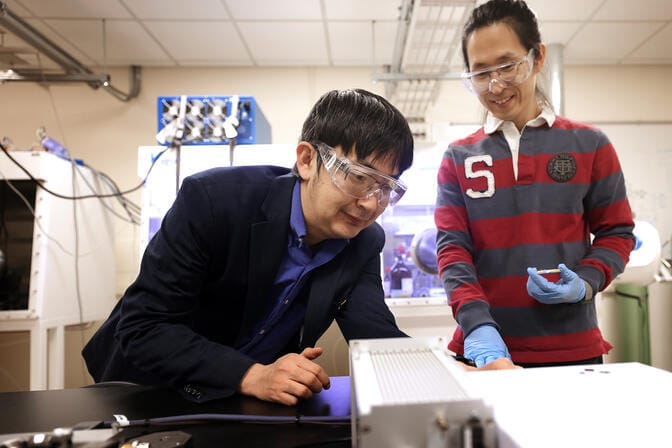 Two researchers are moving the dials on two metal boxes atop a black lab bench.