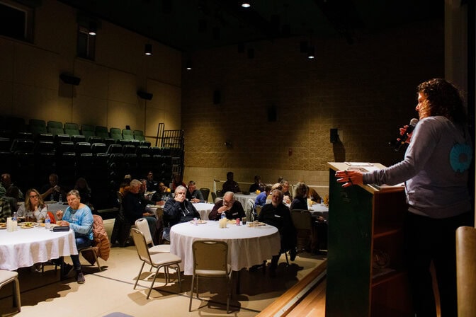  A woman stands at a podium on a stage overlooking seven round tables scattered around a dimly lit gym. Three to seven people sit at each table, which are covered with white table cloths and have candles as a centerpiece.
