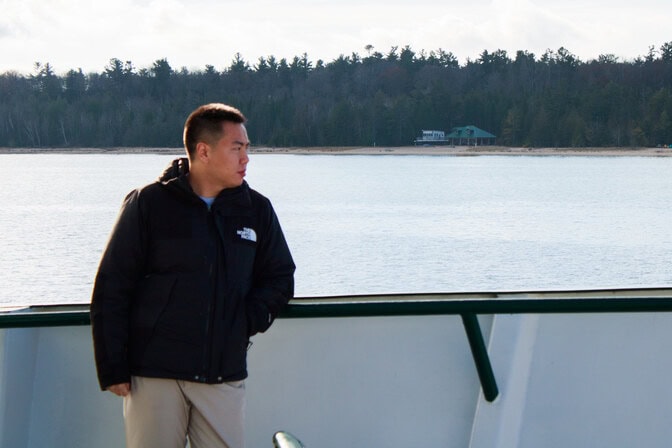 A man leans against the side of a boat, eyes pointed to the horizon. The shoreline of an island is visible behind the man.