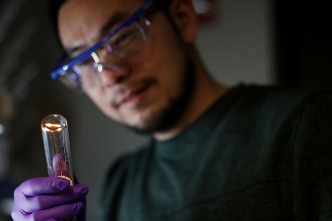  A filament glows golden at the end of a glass tube in Lu's gloved hand. He wears safety glasses.