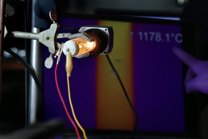 Held in a metal clamp, the bulb glows golden with yellow and red wires attached to the rear. A gloved finger points to a temperature reading of 1178.1 degrees Celsius on the screen behind.