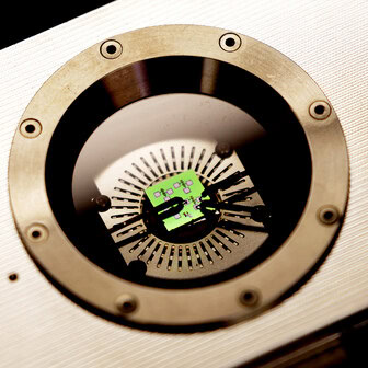 Black pins secure a green computer chip onto a metal platform inside a metal box. A circular window provides a view inside.