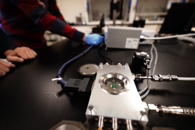 The heater looks like a roughly smartphone-sized metal box with a circular window that allows the researchers to view the green memory device inside. Cables connect the heater to several other metal boxes further down the lab bench.