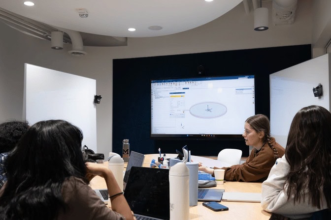 Four young women work on laptops together at a round table. One woman gestures at her laptop screen, which is also being displayed on a large monitor mounted to the wall behind the table. The monitor shows a 3D model of a disc-like part.