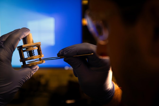 A man holds a thin disc in his right hand with forceps. He inserts the disc into a round clamp, which he holds in his left hand.