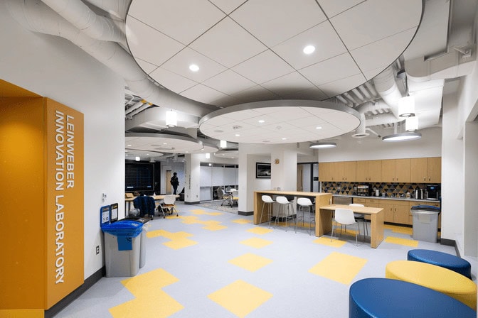 A series of five large, circular discs hang from the ceiling. Each disc has four spotlights that illuminate the lab's yellow and blue-gray tile floor. "Leinweber Innovation Lab" is printed on a section of the wall painted in bright orange.