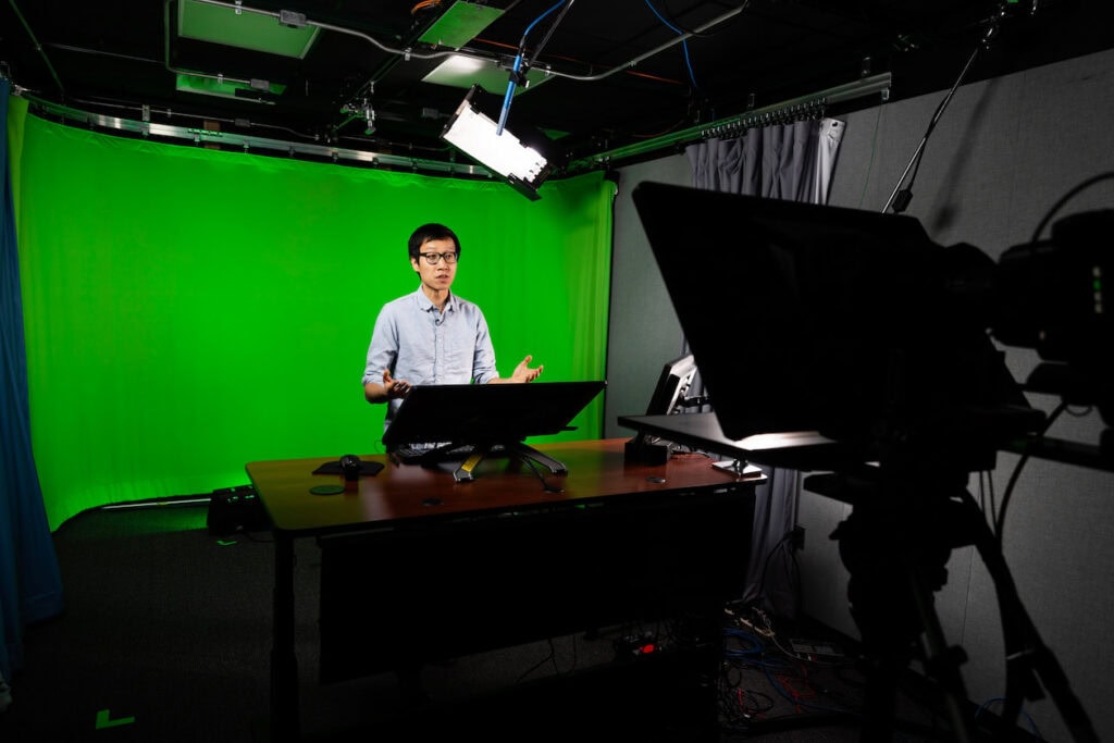 Man in a studio in front of a green screen, speaking and gesturing, with camera and monitors in the foreground.