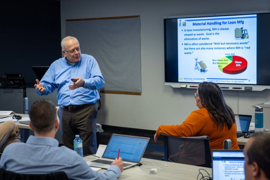 A man gives a presentation on material handling for Lean manufacturing to a group in a classroom setting, with slides displayed on a screen.