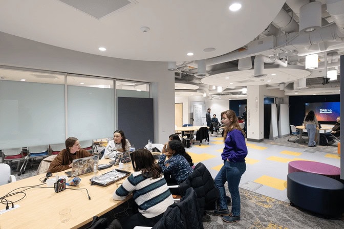 Four young women sit at a round table and work on their laptops. Another woman listens in as she stands near the head of the table. Several other students are working in two additional studios on the opposite side of the lab.