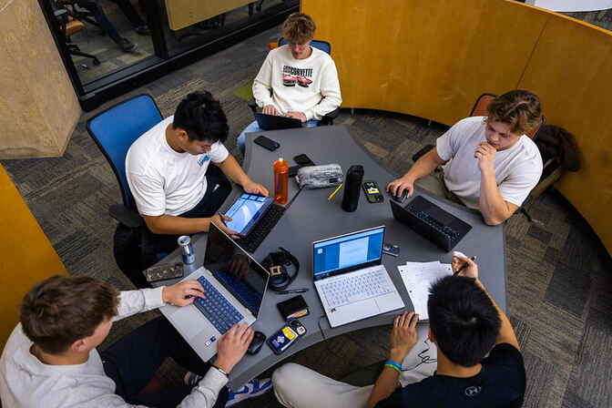 A group of five college students sit around a round table on laptops while engaged in conversation, playing video games together, and studying for final exams.