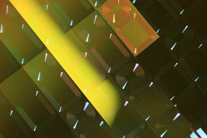 Lines creased into a sheet of gold-colored material mark where individual computer chips will be cut. Each rectangular chip has several channels arranged into a series of two concentric rings. The sheet's color shifts from gold to black as it reflects the factory light.