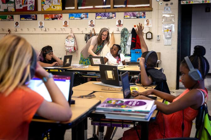 A teacher has walked around an island of four desks to help one of the students sitting at them. The teacher leans over the student's shoulder to point at their laptop screen.
