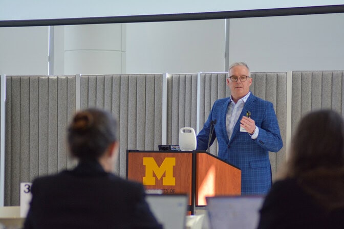 Gregory Salisbury speaking at a podium with the University of Michigan block "M" logo on the front.