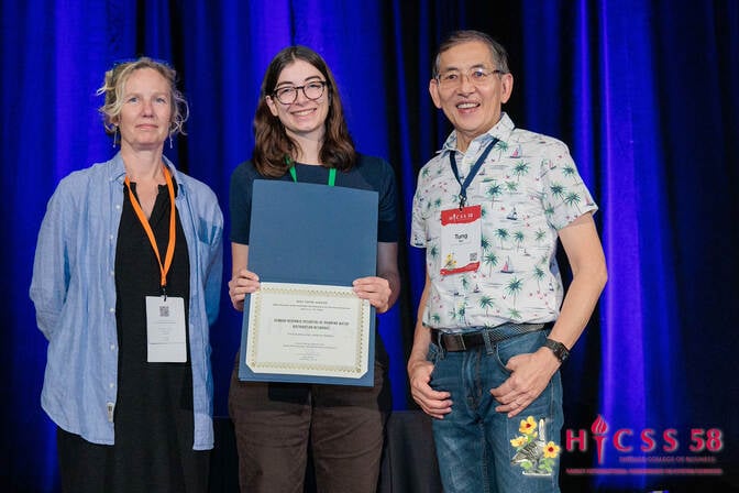 Two women and a man stand on a stage, shoulder to shoulder, smiling. The woman in the middle holds a certificate.