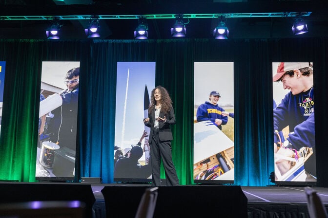 Reitz stands with tall panels featuring scenes from the x88 courses, including the rocket and model plane, behind her. A backdrop of velvet curtains are lit in blue and green. Halow is out of the frame.