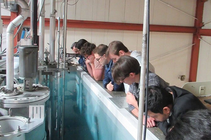 Several people stand side by side along a railing, peering down into a deep water tank filled with clear blue water. The setting is indoors with metal beams, pipes, and equipment around the tank.