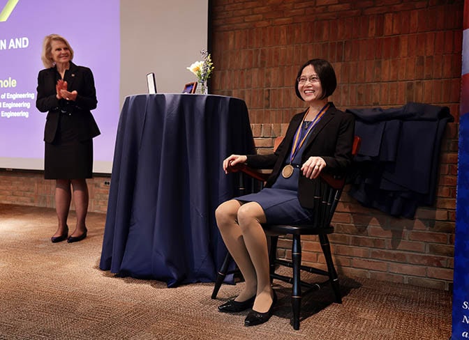 A woman smiles, sitting in a dark stained wooden chair with gold accents, wearing a large medal, while another woman applauds in the background.