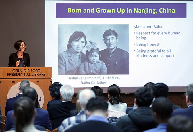 A woman speaks from a podium. The slide reads "Born and Grown Up in Nanjing, China. Mama and Baba: Respect for every human being, Being honest, Being grateful to all kindness and support."

A family photo features Huifen Zhang (mother), Naihu Xu (father), and Little Zhen.