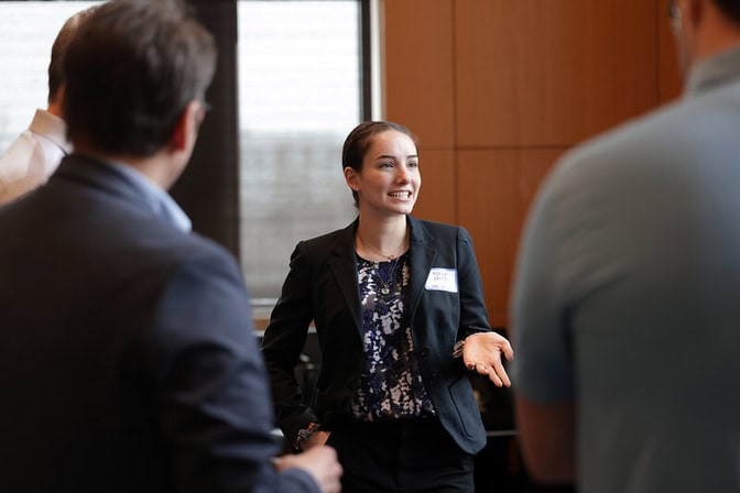 A young woman in a suit speaks with mentors in varying business attire in a wood-paneled conference room.