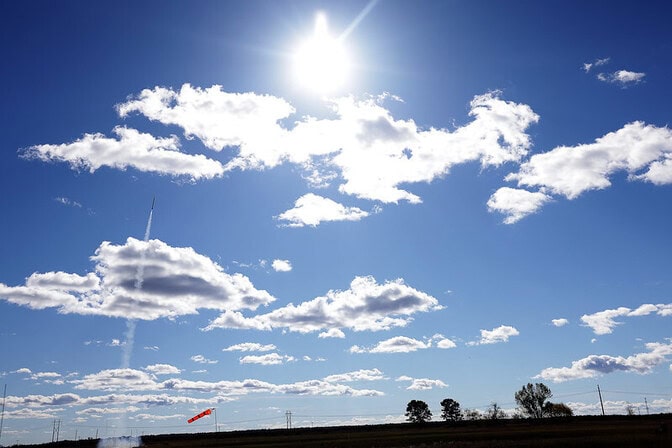 A rocket launches into the sunshine toward the fluffy white clouds.
