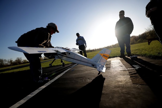 A young man silhouetted against the sun lays the wheels of a large model plane down on a runway while two of his teammates look on.