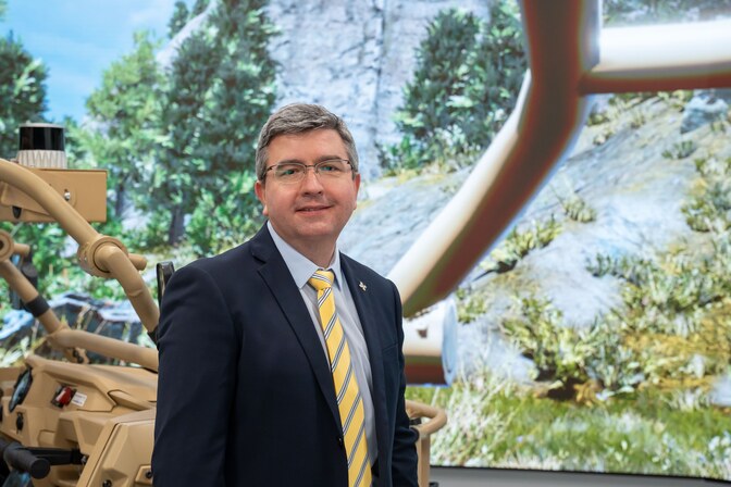 A man stands in front of a wraparound screen displaying a virtual rocky landscape with trees. Part of the roll cage from the physical MRZR is also visible behind him.