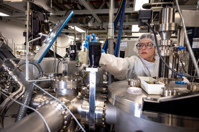 Claire Arneson in a lab coat, hairnet and safety glasses, reaches over a large metal contraption with many tubes and pipes.